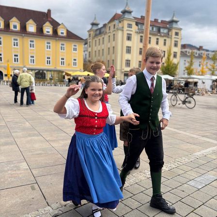 Ein Junge und ein Mädchen gehen lächelnd auf einem Platz mit Gebäuden im Hintergrund.