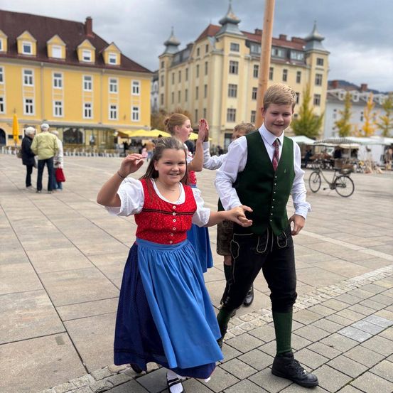 Ein Junge und ein Mädchen gehen lächelnd auf einem Platz mit Gebäuden im Hintergrund.
