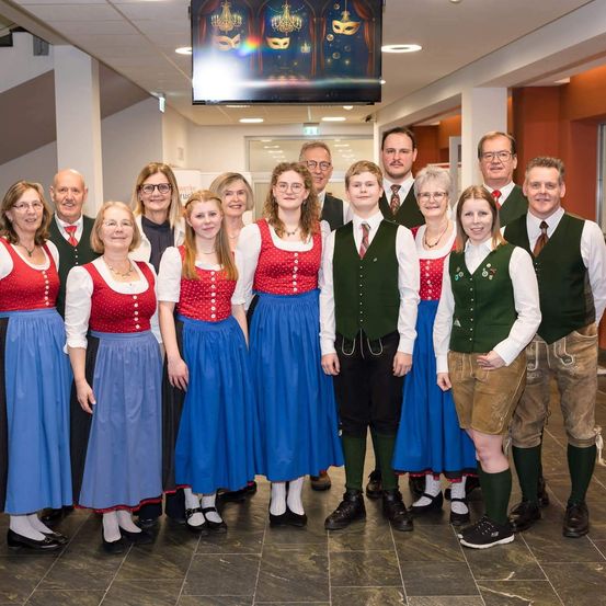 Eine Gruppe von Menschen in traditioneller Kleidung posiert für ein Foto in einem Raum. Einige tragen Westen und Krawatten, andere Röcke und Blusen. Eine große Leinwand zeigt ein farbenfrohes Bild.