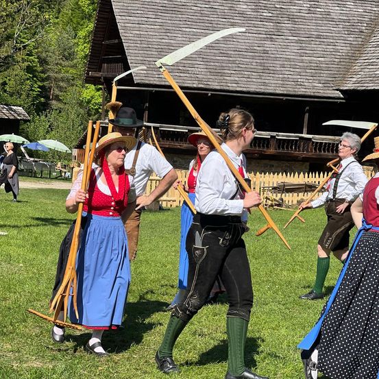 Eine Gruppe von Menschen in traditioneller bayerischer Kleidung geht auf einem Rasenfeld mit holzernen Werkzeugen, mit einem rustikalen Gebäude im Hintergrund.