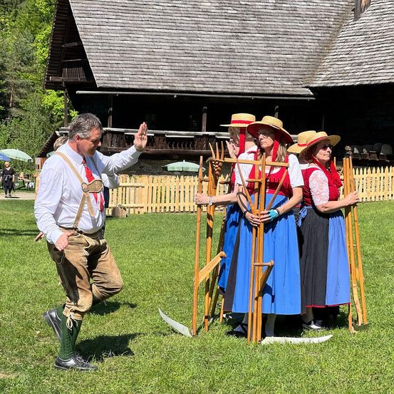 Ein Mann in traditioneller Kleidung winkt einer Gruppe von Frauen in gleichen Outfits zu, die Holzwerkzeuge halten. Sie stehen vor einem Holzzaun mit einem Haus im Hintergrund.