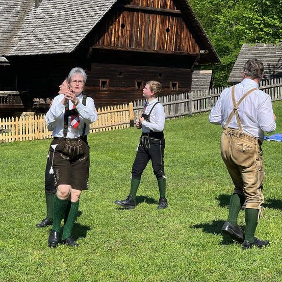 Drei Personen in traditioneller Kleidung stehen auf einem Rasen. Eine Frau hält eine Maske. Hinter ihnen steht ein Holzhaus mit einem Zaun.