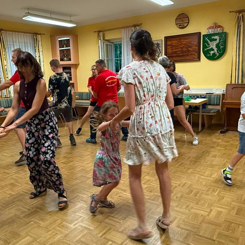 A group of people are dancing in a room with wooden floors and yellow walls. A young girl in a floral dress is being led by a woman.
