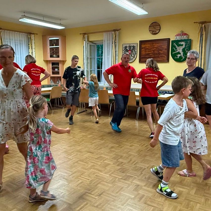 A group of people, including adults and children, are dancing in a room with wooden floors and yellow walls. Some are wearing red shirts with text, while others are in casual clothing. There are chairs and tables in the background.