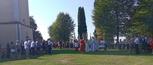 Eine Gruppe von Menschen versammelt sich in einem Park für eine Zeremonie, vor einer Kirche mit einigen Bäumen. Einige tragen traditionelle Kleidung.