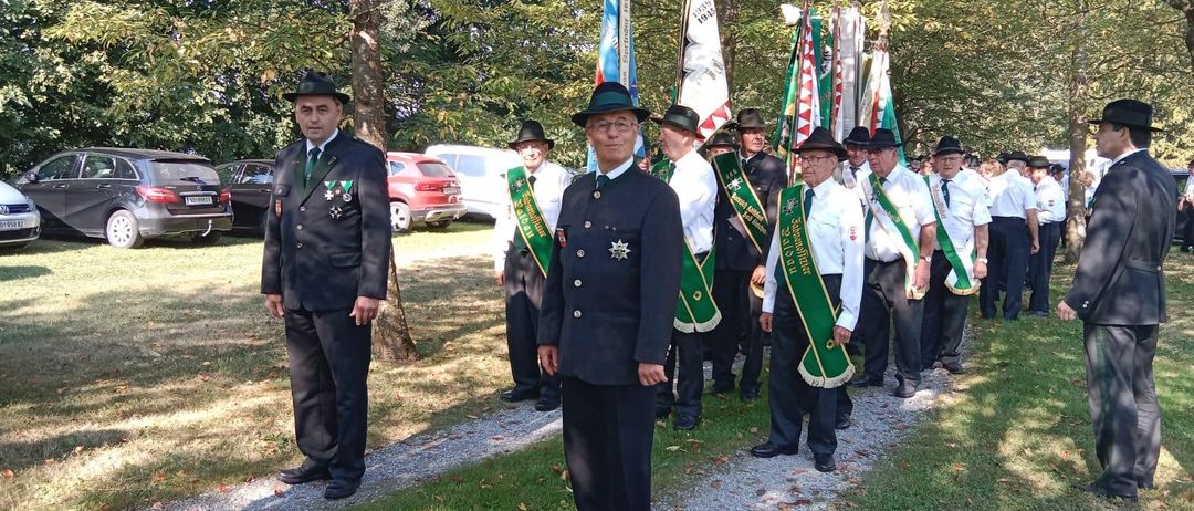 Eine Gruppe uniformierter Männer, möglicherweise eine Musikkapelle, steht auf einem Kiesweg mit Fahnen hinter ihnen. Sie befinden sich in einem Park mit Bäumen und geparkten Autos.