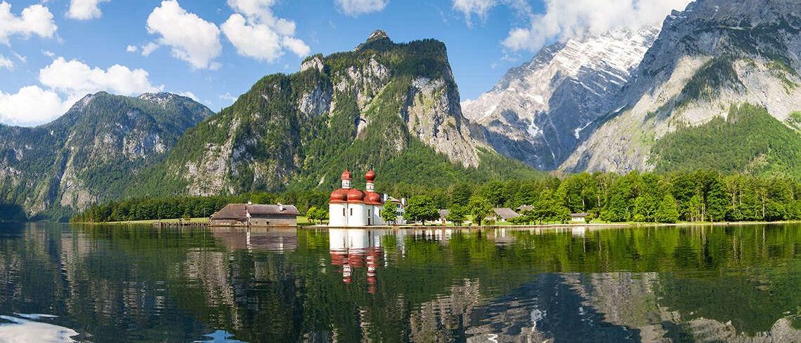 Ein ruhiger See mit einer kleinen Kapelle in der Mitte, umgeben von majestätischen Bergen und einem blauen Himmel mit Wolken.