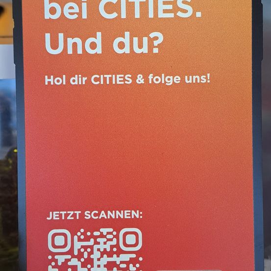 Ein rotes Schild mit weißem Text lautet 'bei CITIES. Und du? Hol dir CITIES & folge uns!' und einem QR-Code.
