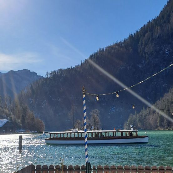 Eine Fähre fährt auf einem See mit Bergen und Bäumen im Hintergrund. Ein blau-weißer Pfahl ist im Vordergrund.