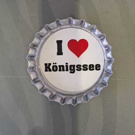 Eine metallische Kappe mit den Worten 'I love Konigssee' darauf. Der Buchstabe 'I' ist schwarz, gefolgt von einem roten Herzen und 'love' in Schwarz. 'Konigssee' steht darunter in schwarz.