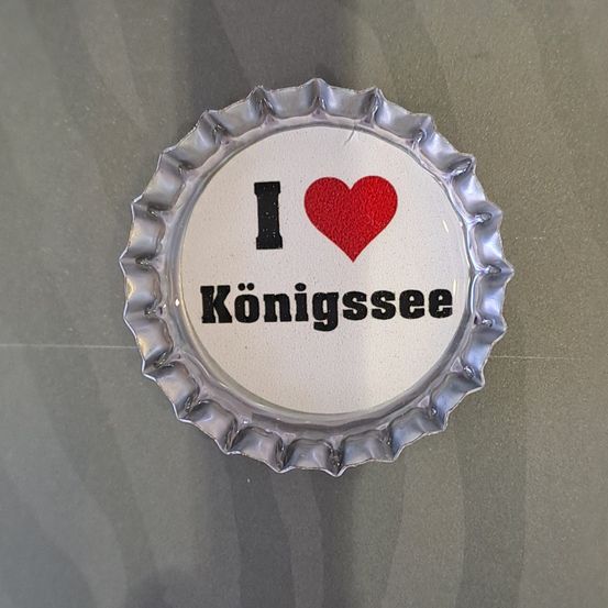 Eine metallische Kappe mit den Worten 'I love Konigssee' darauf. Der Buchstabe 'I' ist schwarz, gefolgt von einem roten Herzen und 'love' in Schwarz. 'Konigssee' steht darunter in schwarz.