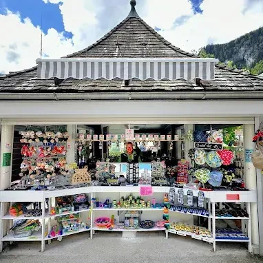 Ein kleiner Laden mit einem kegelförmigen Dach ist mit verschiedenen Artikeln gefüllt, darunter Hüte und Taschen. Eine Person ist drinnen, und Berge sind im Hintergrund.