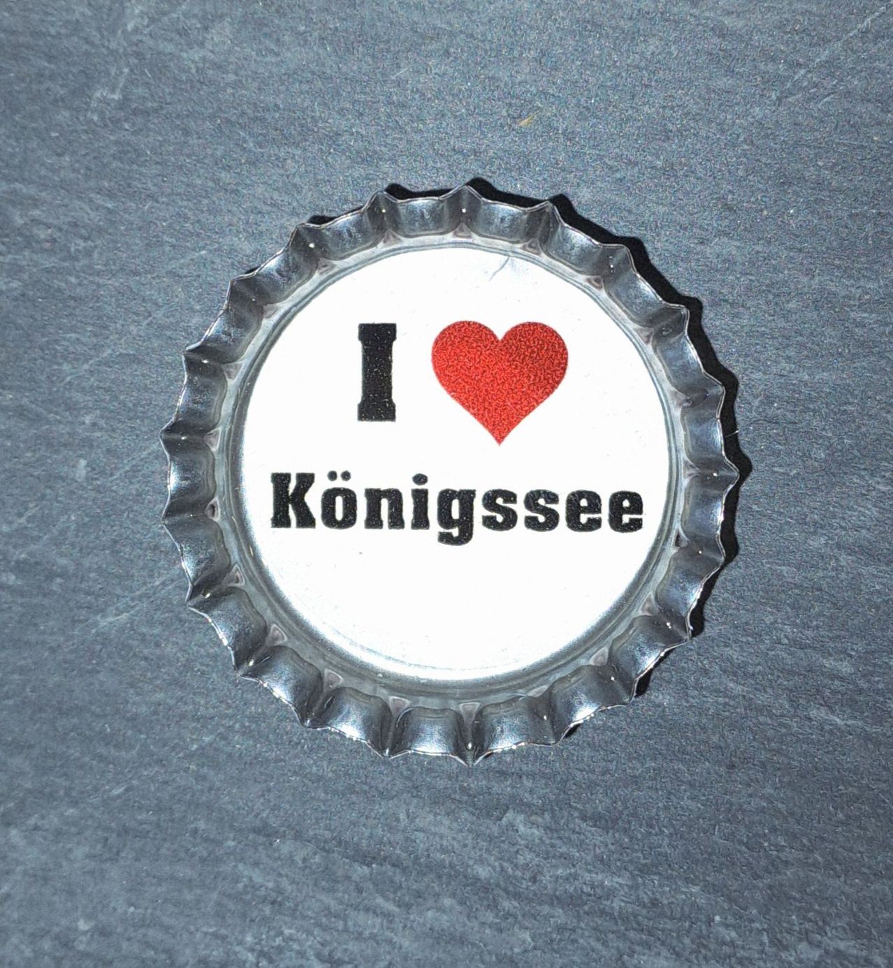 Eine Metallkappe mit weißem Hintergrund, auf der die Worte 'Ich liebe Königssee' und ein rotes Herz in der Mitte stehen.