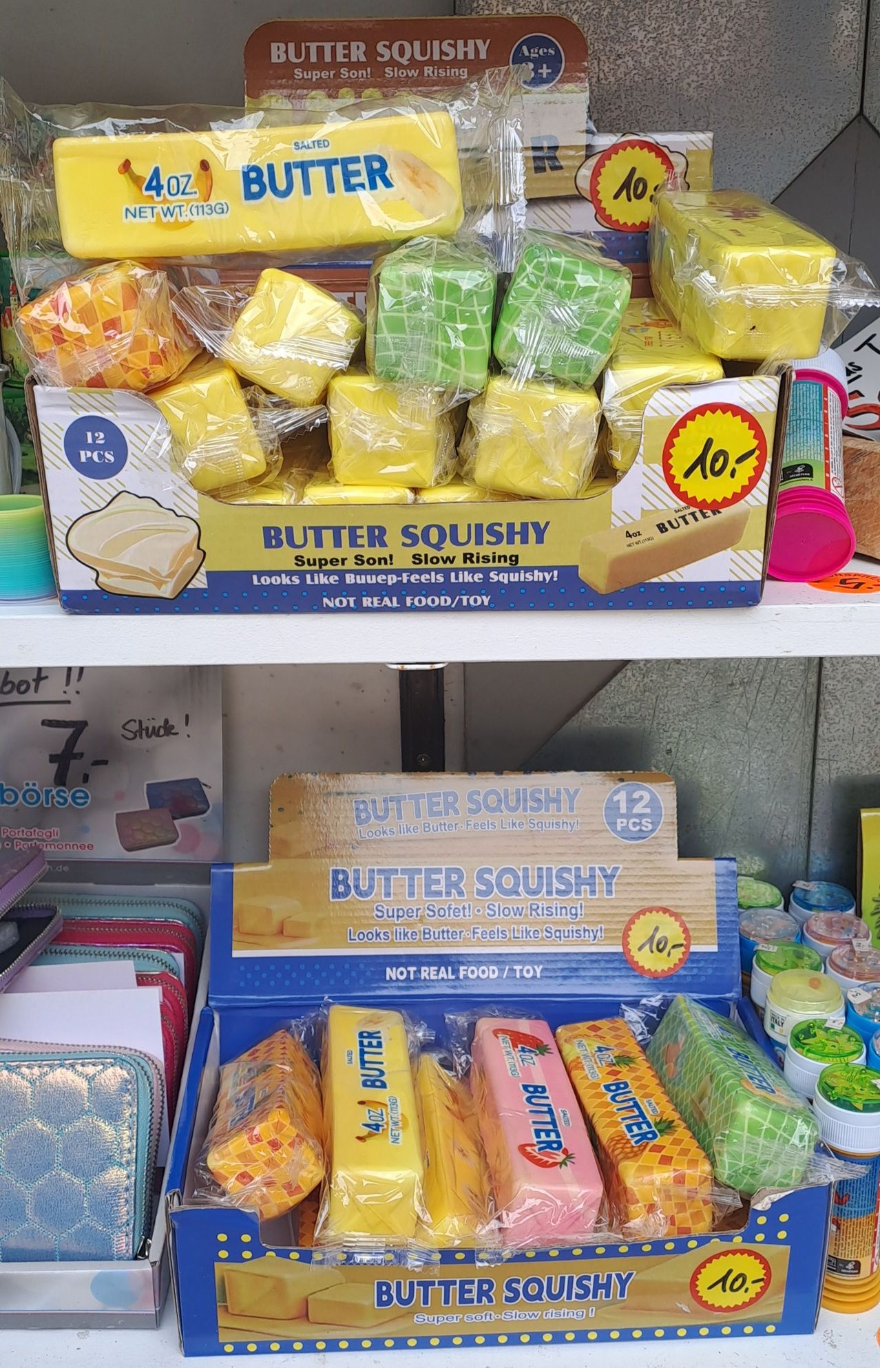 Eine Box mit Butter Squishy Spielzeug auf einem Regal mit anderen Artikeln, einschließlich eines rosa Deckels und einer blauen Box mit gelbem Deckel.