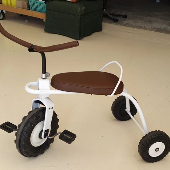 Ein Dreirad mit braunem Sitz und schwarzen Reifen steht auf einem weißen Boden. Es hat einen braunen Lenker und Fußpedale.