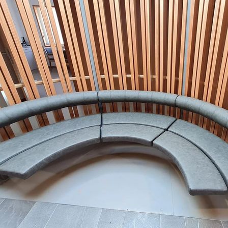 Eine gewinkelte graue Ledercouch steht vor einer Holzwand. Die Couch hat mehrere Kissen und scheint sich in einer modernen Inneneinrichtung zu befinden.