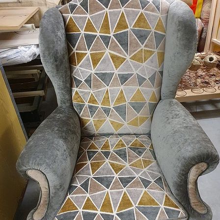Ein grauer Ohrensessel mit einem geometrischen Muster in Beige, Gold und Grau. Der Sessel ist aus grauem Stoff gefertigt und hat einen Holzfuß. Hinter dem Sessel befindet sich ein Schreibtisch mit einer Holzplatte und einer Stoffrolle.