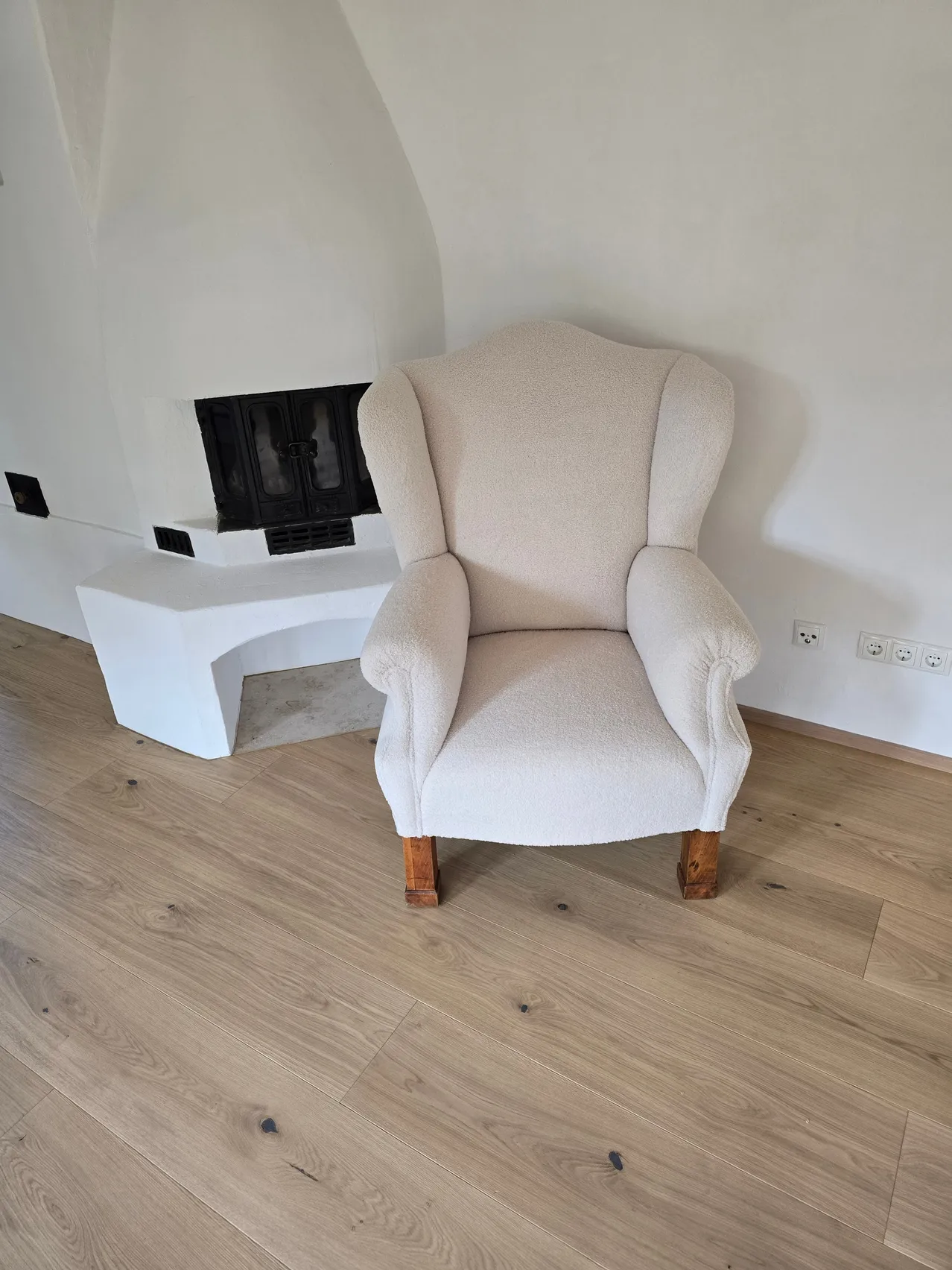 Ein weißer Sessel mit Holzbeinen steht in einem Raum mit Kamin und weißer Wand. Der Boden ist aus Holz.