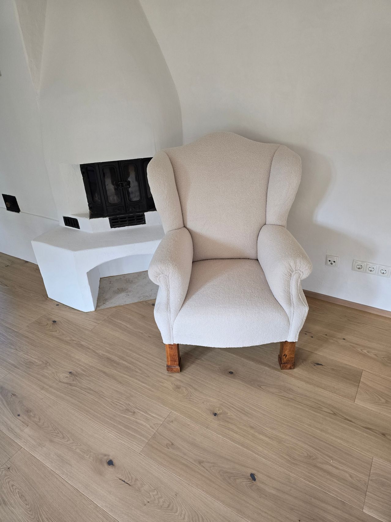Ein weißer Sessel mit Holzbeinen steht in einem Raum mit Kamin und weißer Wand. Der Boden ist aus Holz.