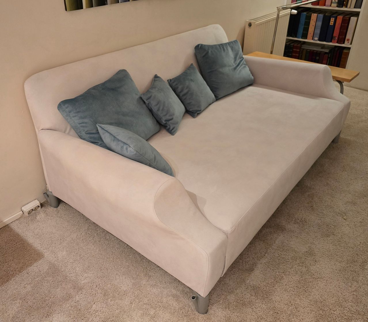 Ein weißer Sofa mit beigefarbenem Bezug, ausgestattet mit vier blauen Kissen, auf einem Teppichboden platziert, mit einem Bücherregal und einem Heizkörper im Hintergrund.