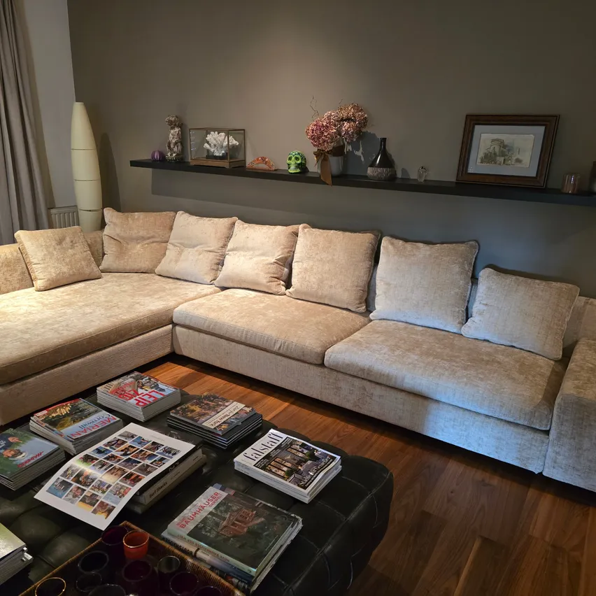 Ein Wohnzimmer mit einem beigen Ecksofa, mehreren Kissen und einem Holzboden. Zeitschriften und Bücher liegen auf einem Couchtisch. Ein Regal mit verschiedenen Gegenständen befindet sich über dem Sofa.