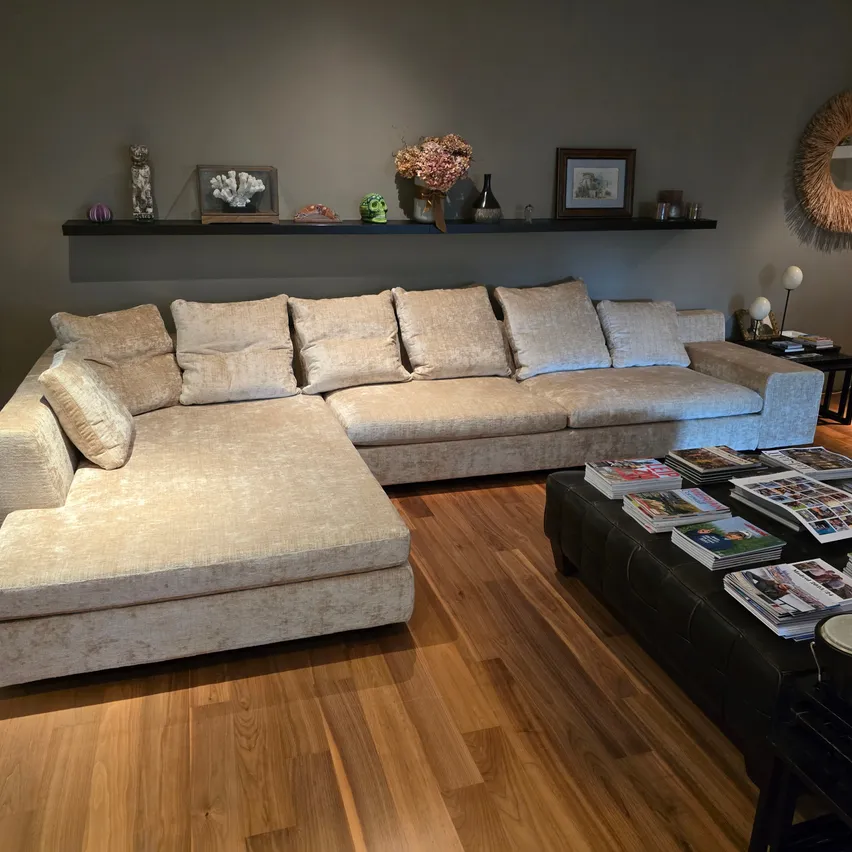 Ein beiger Ecksofa mit Kissen befindet sich in einem Wohnzimmer. Es steht auf einem Holzboden. Es gibt einen schwarzen Couchtisch mit Zeitschriften. Ein Regal darüber hat eine Vase und andere Gegenstände.
