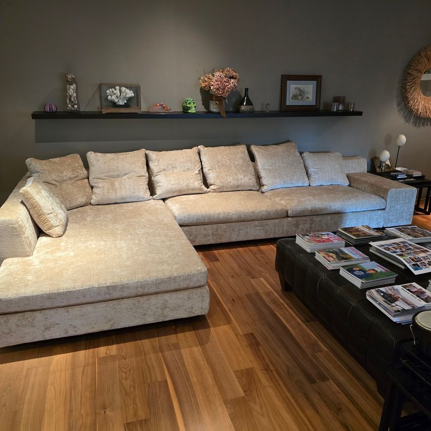 Ein beiger Ecksofa mit Kissen befindet sich in einem Wohnzimmer. Es steht auf einem Holzboden. Es gibt einen schwarzen Couchtisch mit Zeitschriften. Ein Regal darüber hat eine Vase und andere Gegenstände.