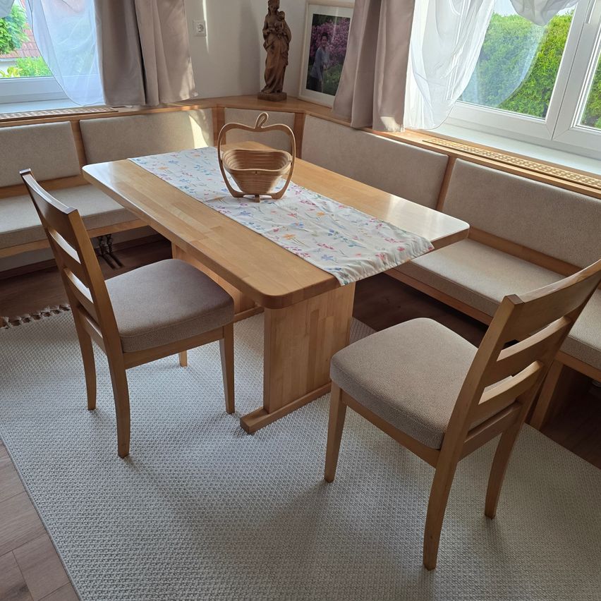 Ein Eck-Essbereich mit einem Holztisch und Stühlen, einem Korb mit einem Apfel und einer Bank mit Kissen. Eine Statue und ein Bilderrahmen befinden sich über der Bank. Der Raum hat weiße Vorhänge und Glasfenster.