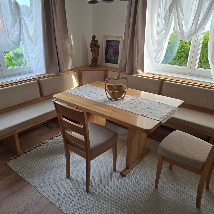 Eine Eck-Essecke mit einem holzfarbenen Tisch und Stühlen, einem Korb mit einem Apfel und einer Statue in einer Ecke. Der Raum hat zwei Fenster mit weißen Vorhängen.