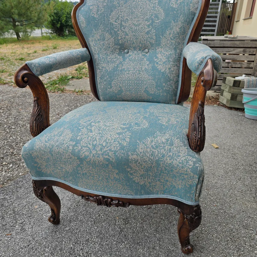 Ein verzierter Sessel mit blauem Stoffbezug und hölzernen Armlehnen steht auf einem Kiesweg. Dahinter sind ein Holzlager und eine Leiter zu sehen.