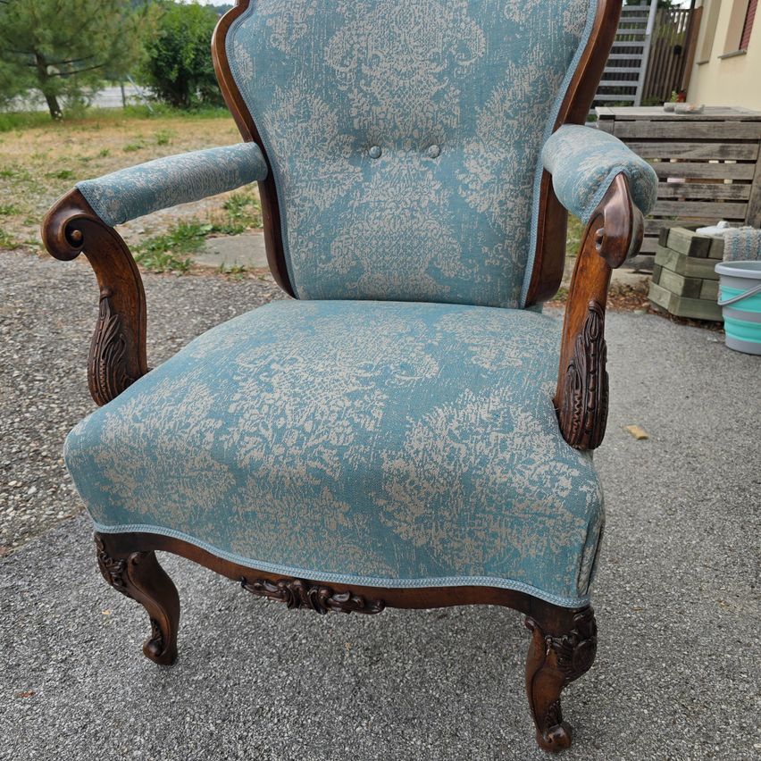 Ein verzierter Sessel mit blauem Stoffbezug und hölzernen Armlehnen steht auf einem Kiesweg. Dahinter sind ein Holzlager und eine Leiter zu sehen.