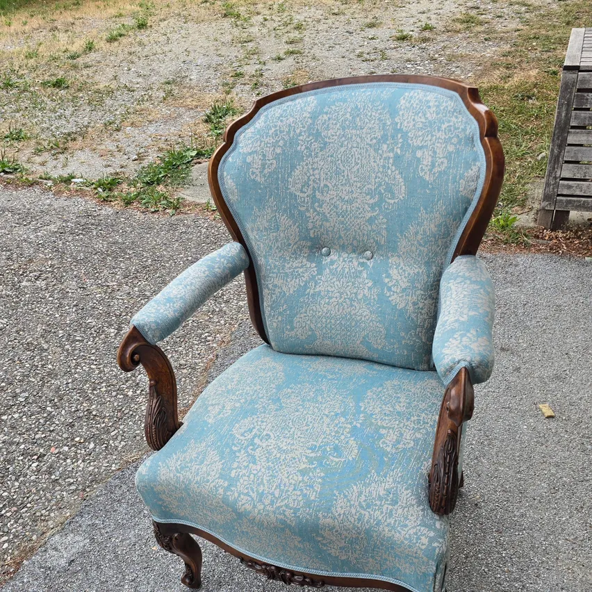 Ein antiker Sessel mit blauer Polsterung und braunem Holzrahmen steht auf einem Schotterweg. Der Sessel hat ein aufwendiges Design auf der Polsterung.