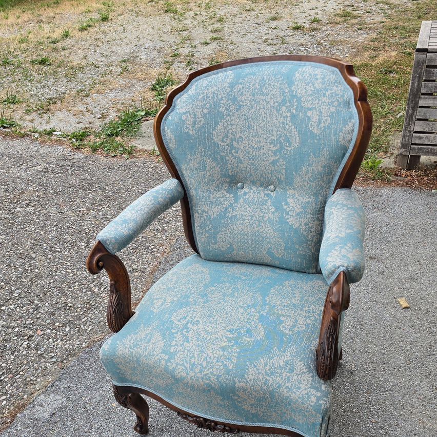 Ein antiker Sessel mit blauer Polsterung und braunem Holzrahmen steht auf einem Schotterweg. Der Sessel hat ein aufwendiges Design auf der Polsterung.