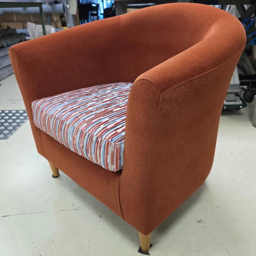 Ein orangefarbener Sessel mit einem gestreiften Kissen ist in einer Werkstatt zu sehen. Der Stuhl hat Holzbeine und eine glatte Stoffoberfläche.