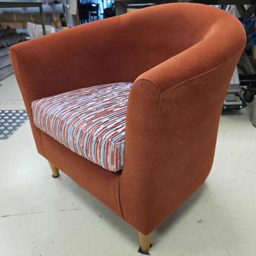 Ein orangefarbener Sessel mit einem gestreiften Kissen ist in einer Werkstatt zu sehen. Der Stuhl hat Holzbeine und eine glatte Stoffoberfläche.