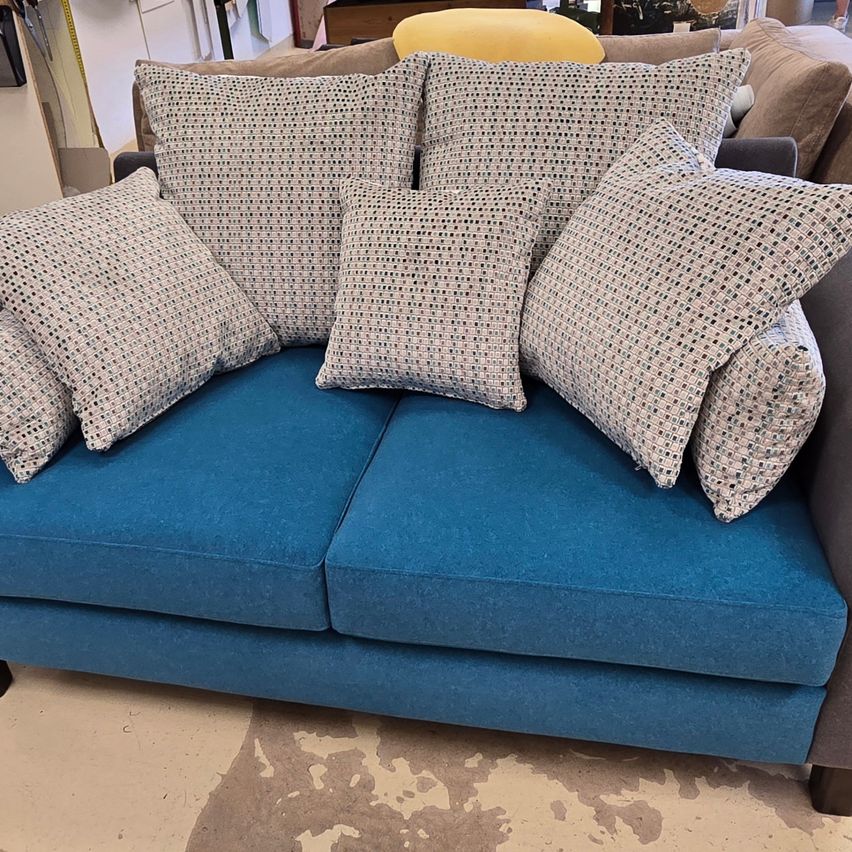 Eine blaue Couch mit mehreren Kissen, darunter ein großes, in einem Raum mit weißer Wand und einem gelben Stuhl.