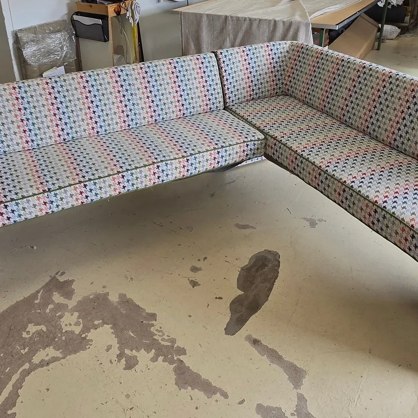 Ein farbenfrohes Sofa mit einem Dackelmuster steht an einer Wand, neben einem abgedeckten Tisch und verschiedenen Gegenständen. Der Boden weist Abnutzungsspuren auf.