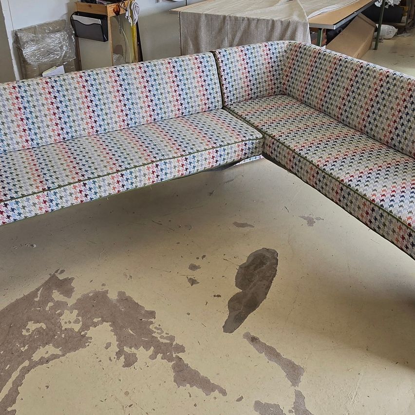 Ein farbenfrohes Sofa mit einem Dackelmuster steht an einer Wand, neben einem abgedeckten Tisch und verschiedenen Gegenständen. Der Boden weist Abnutzungsspuren auf.