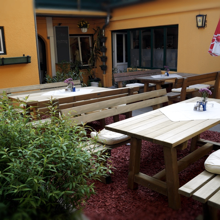 Terrassenbereich mit Holztischen und Bänken. Blumentöpfe, eine geschlossene Tür und ein roter Schirm sind vorhanden.