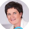 Eine lächelnde Frau mit kurzen Haaren, die ein weißes Kleid trägt, steht vor einem weißen Hintergrund mit bunten Kreisen um sie herum.