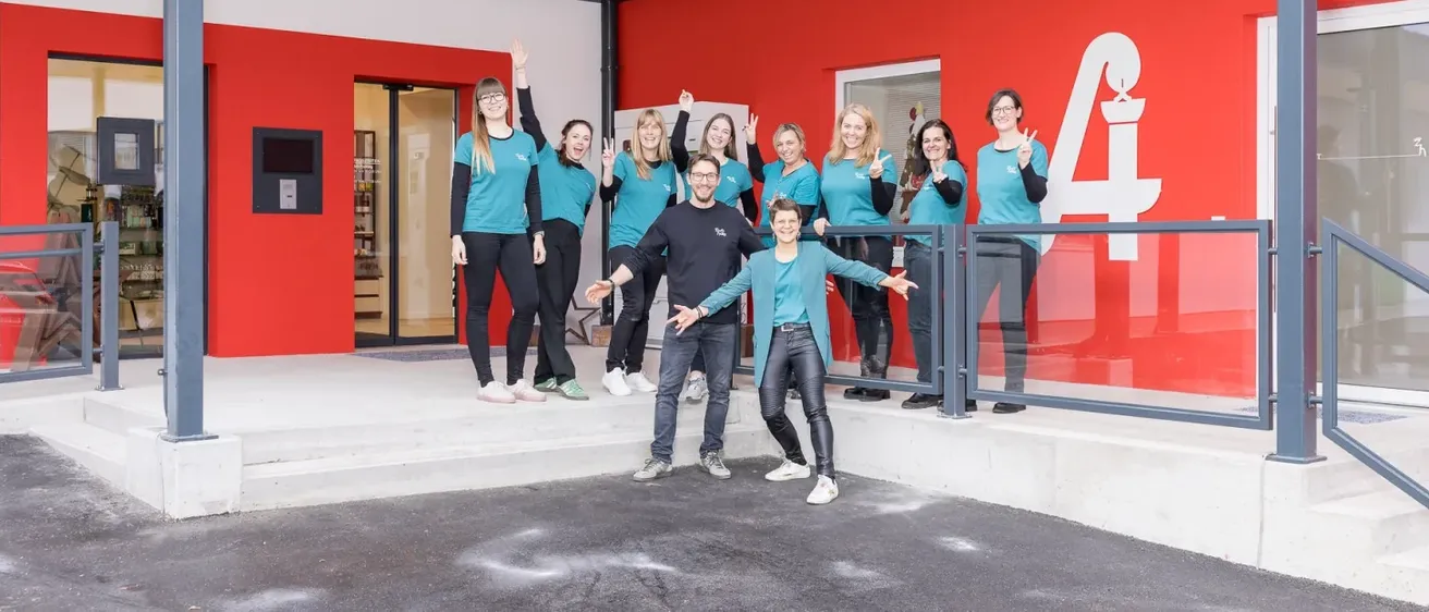 Eine Gruppe von Menschen in passenden blauen T-Shirts und schwarzen Hosen posiert vor einer Apotheke mit einer roten Wand und einem Glaseingang.
