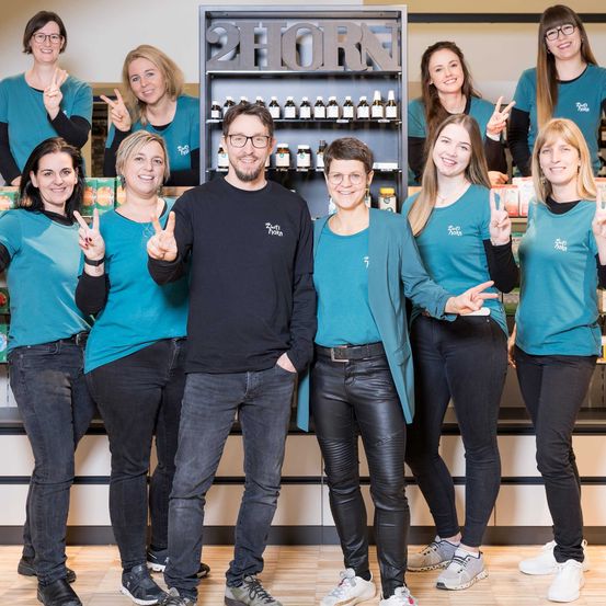 Eine Gruppe lächelnder Personen, sowohl Männer als auch Frauen, steht in einer Reihe und posiert wahrscheinlich für ein Foto. Sie tragen passende blaue Hemden und schwarze Hosen. Hinter ihnen zeigt ein Geschäft verschiedene Produkte an.