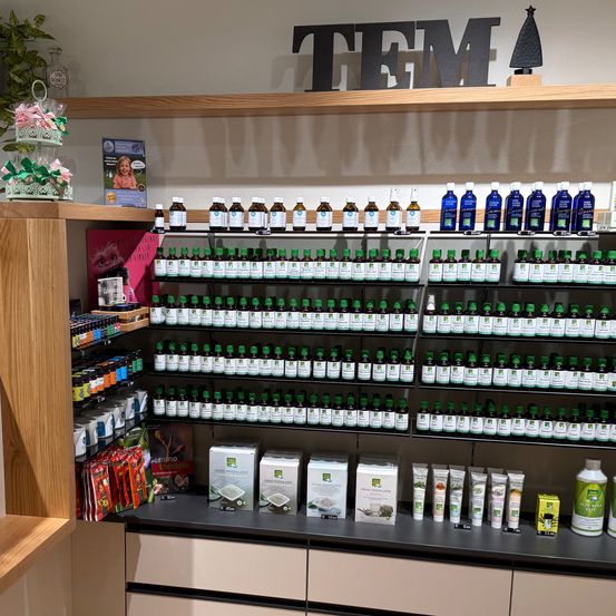 Ein Ladeninnenraum mit Regalen voller grüner Flaschen, beschriftet mit 'TEM'. Eine Theke mit einer Pflanze, einem Schild 'PANIER DES SENS' und verschiedenen Artikeln. Eine Holzbank und ein Holzregal mit Produkten.