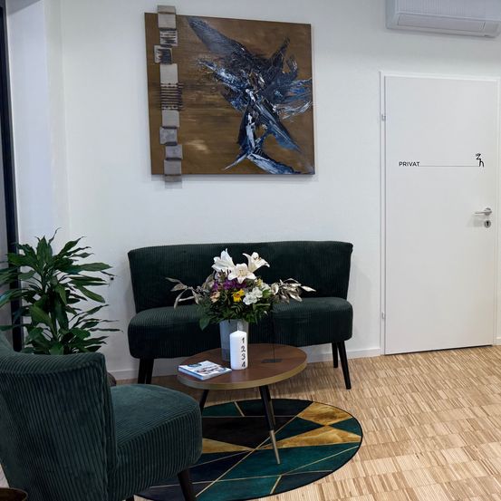 Ein Raum mit Holzböden, weißen Wänden und einer weißen Tür. Ein Sofa mit Blumen, ein runder Tisch und ein Teppich. Ein Displayregal mit Flaschen und Behältern. Ein Schrank mit Pflanzen und einem Gemälde an der Wand.