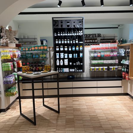 Ein Blick in das Innere eines Ladens zeigt ein großes schwarzes Regal mit der Aufschrift '2HORN', gefüllt mit verschiedenen Produkten, einem Tisch mit Tellern voller Essen und Regalen mit unterschiedlichen Artikeln. Der Boden ist aus Holz.