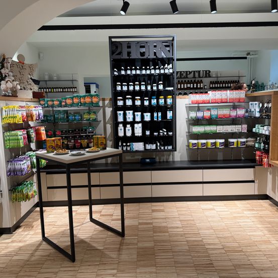Ein Blick in das Innere eines Ladens zeigt ein großes schwarzes Regal mit der Aufschrift '2HORN', gefüllt mit verschiedenen Produkten, einem Tisch mit Tellern voller Essen und Regalen mit unterschiedlichen Artikeln. Der Boden ist aus Holz.