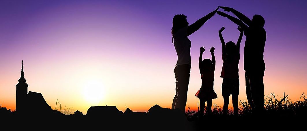 Eine Familie mit vier Personen, zwei Erwachsenen und zwei Kindern, steht in Silhouette vor einem Sonnenuntergang. Sie halten Händchen und schaffen einen berührenden Familienmoment.