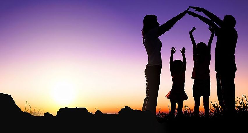 Eine Familie mit vier Personen, zwei Erwachsenen und zwei Kindern, steht in Silhouette vor einem Sonnenuntergang. Sie halten Händchen und schaffen einen berührenden Familienmoment.