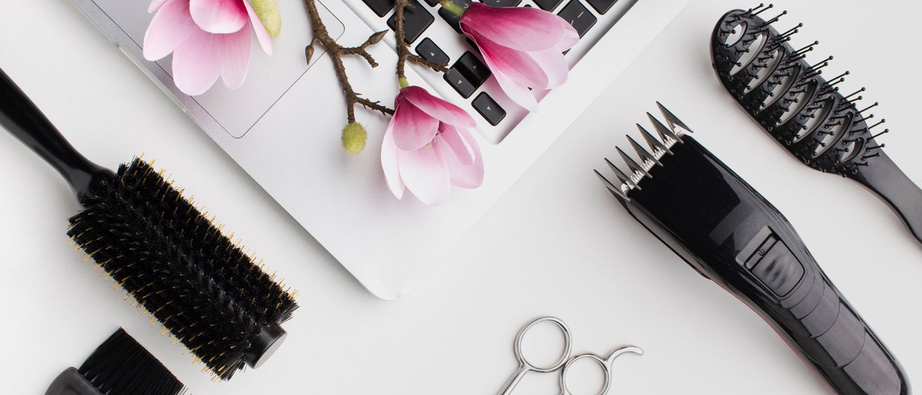 Ein Laptop mit rosa Blumen darauf, ein schwarzer Haarschneider, eine Bürste und Scheren liegen auf einer weißen Oberfläche.