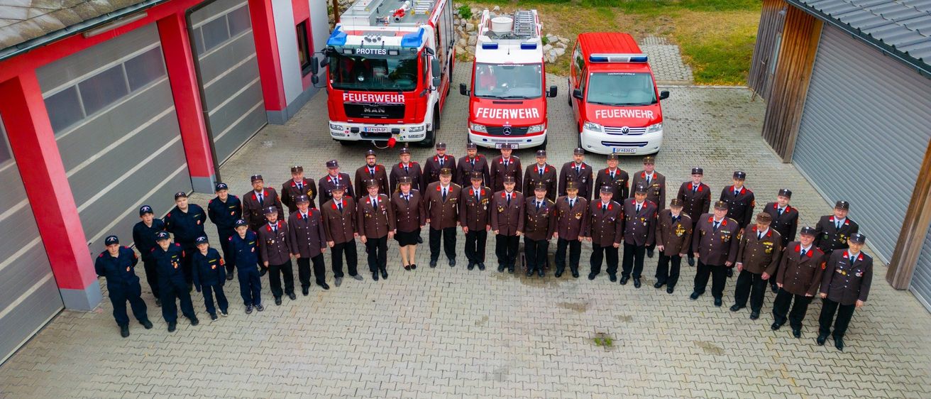 Eine Gruppe Feuerwehrleute in Uniform posiert für ein Foto vor ihren Feuerwehrfahrzeugen. Die Szene spielt sich in einem ländlichen Bereich ab, mit Gebäuden im Hintergrund.
